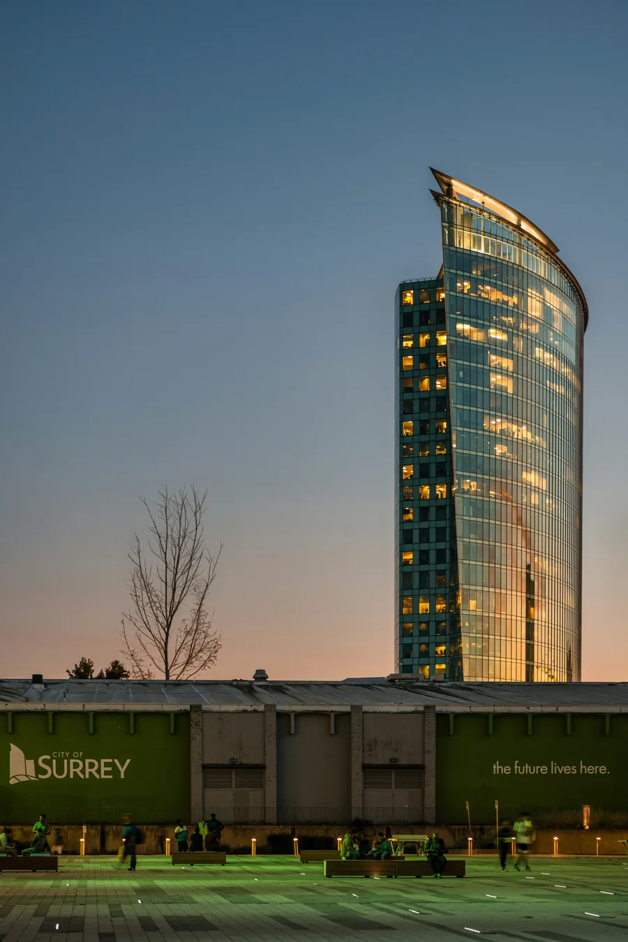 The Surrey Civic Plaza tower lit at dusk, with the Surrey sign in front, near Surrey Central Station